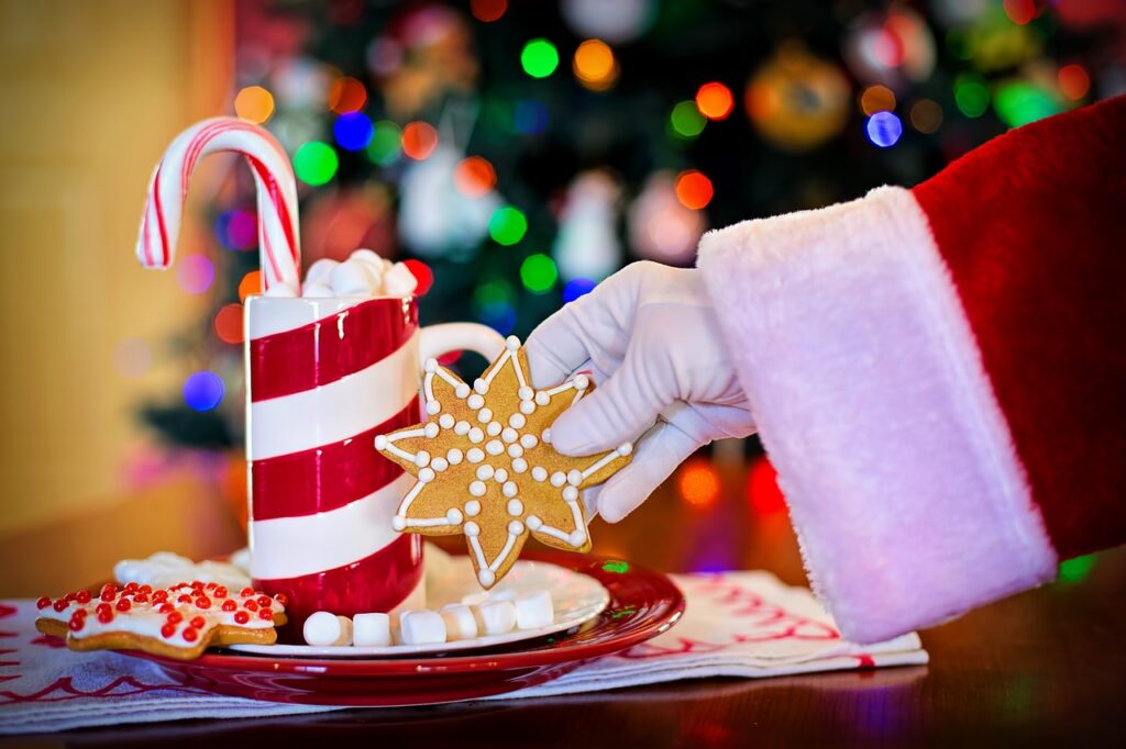 Santa's hand reaching for a festive gingerbread cookie on a candy cane-striped mug surrounded by holiday treats.