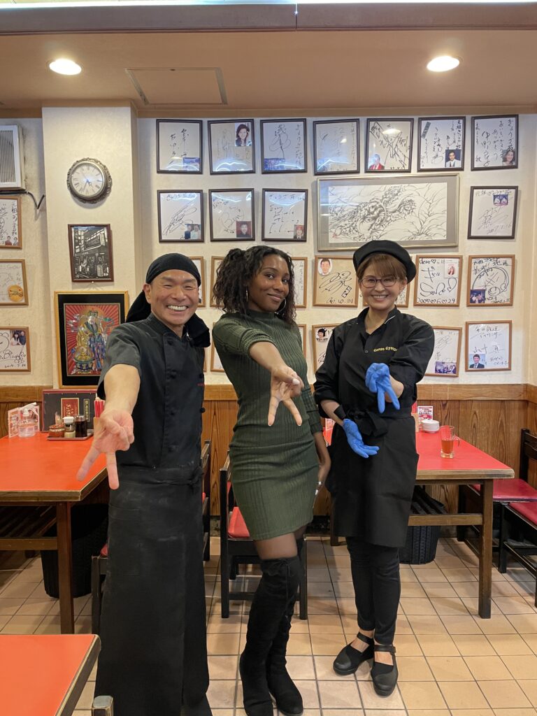 Three people posing together inside a cozy restaurant, with framed autographs adorning the wall behind them.