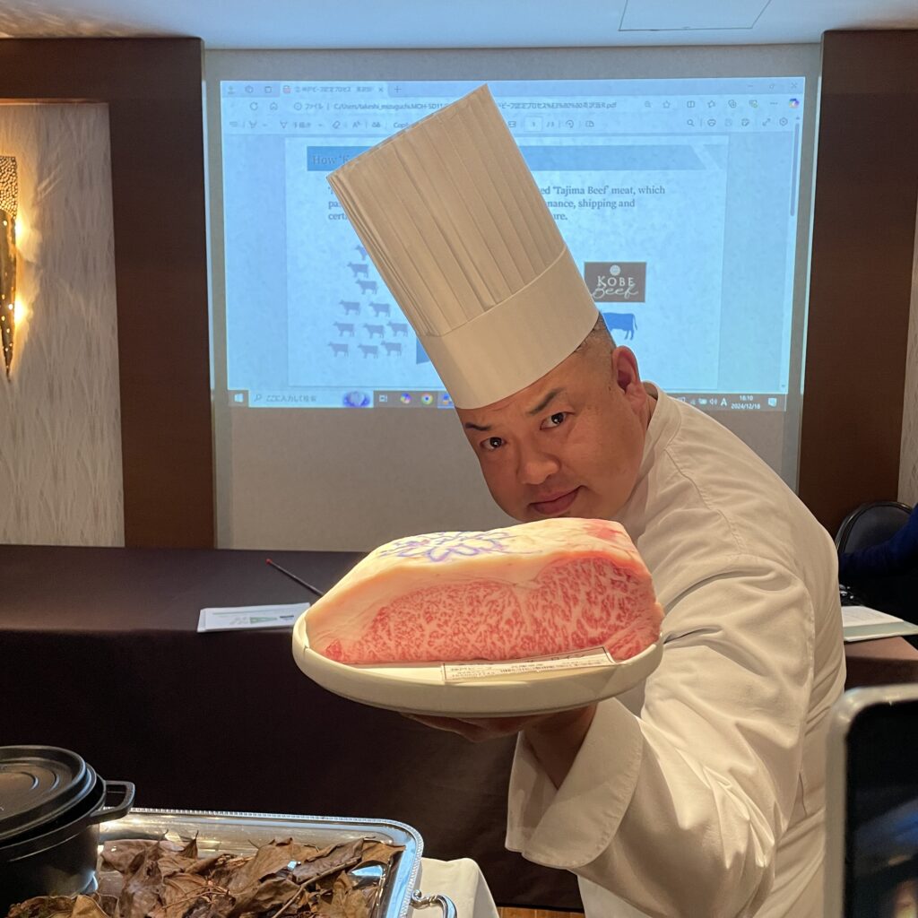 A chef holding a plate with premium Kobe beef on display, wearing a tall white hat and smiling.