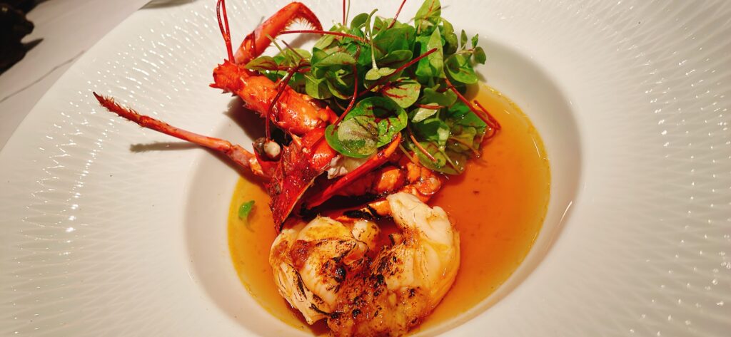  A beautifully plated dish of lobster with greens and a rich broth in a white bowl.