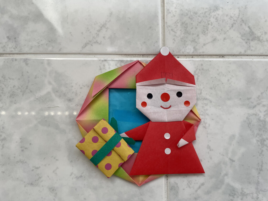 A cute origami Santa Claus holding a colorful gift, placed against a tiled wall.