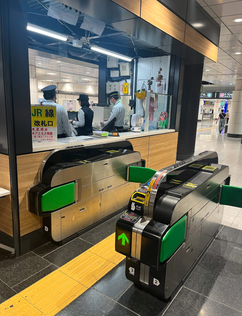 A ticket gate at a JR train station in Tokyo, with uniformed staff assisting travelers at the information counter nearby.