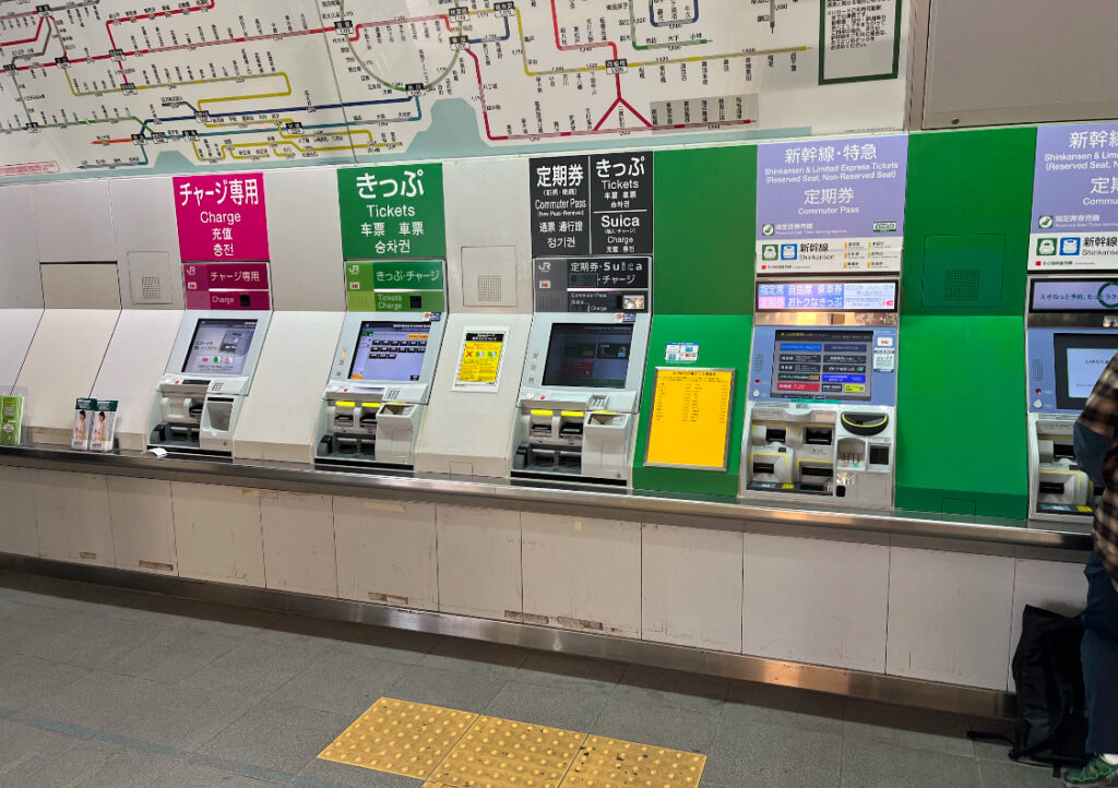  A row of ticket machines in a JR station, labeled with options for ticket purchases, Suica card charging, and commuter passes, with a detailed train map above.