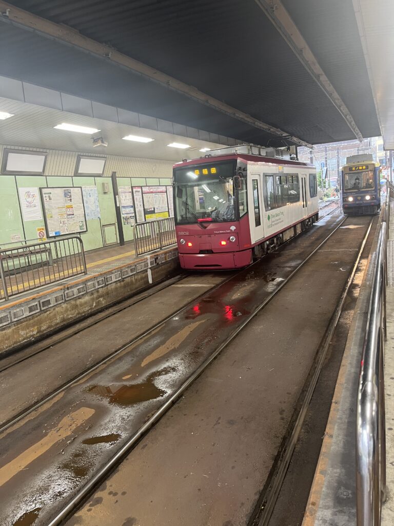 A red Sakura Tram arriving at Otsuka Station, with tracks glistening from rain and a covered waiting platform in view.