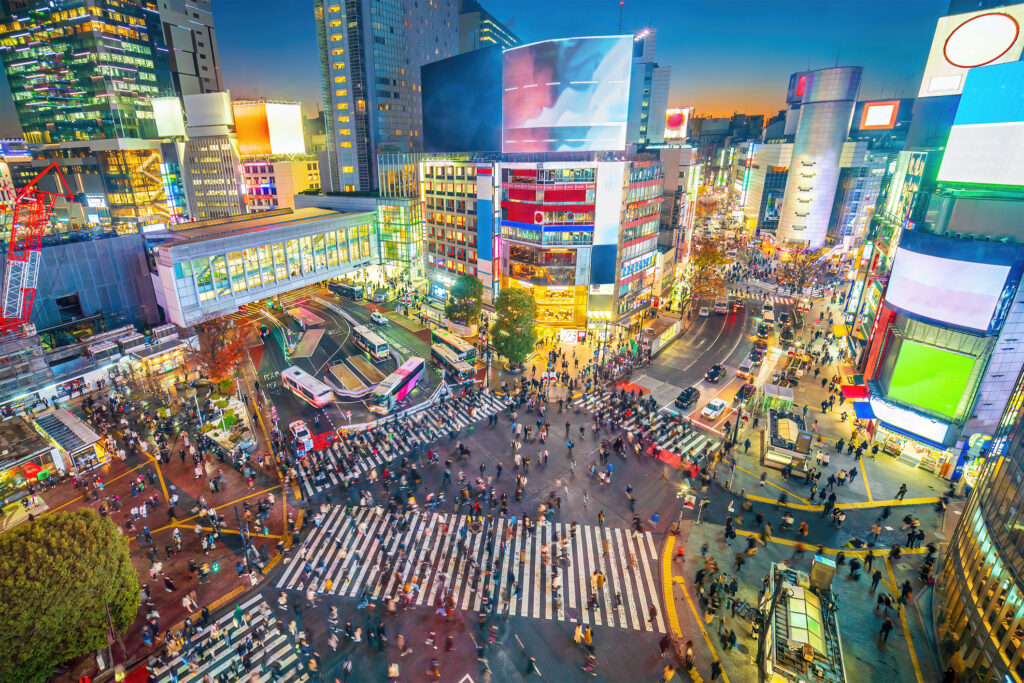 A vibrant view of Shibuya Crossing at sunset with crowds of people, illuminated buildings, and busy streets in Tokyo, Japan.