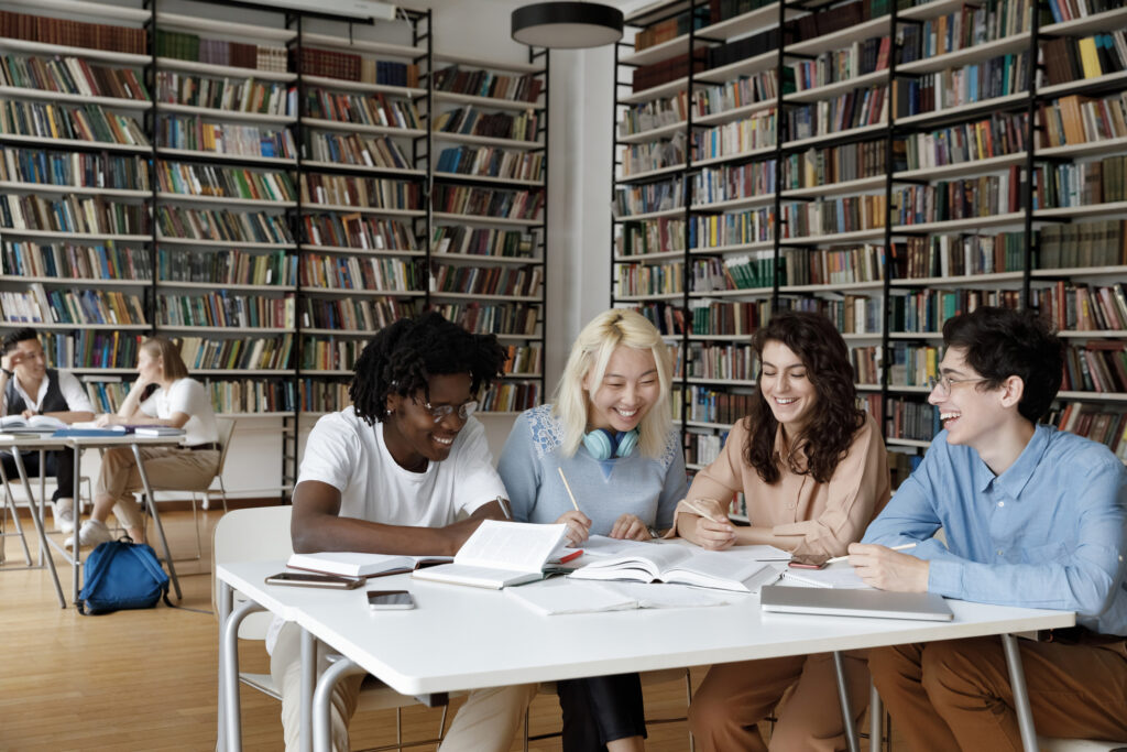 A diverse group of international students studying together at a table in a library, with bookshelves in the background.