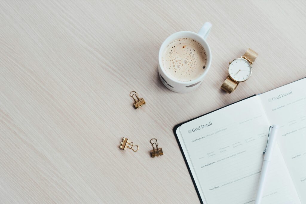 A cup of coffee, a watch, and an open planner on a wooden desk.