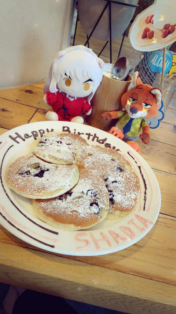 A plate of blueberry pancakes with "Happy Birthday Shadia" written in chocolate and pink icing, accompanied by plush toys on a wooden table.