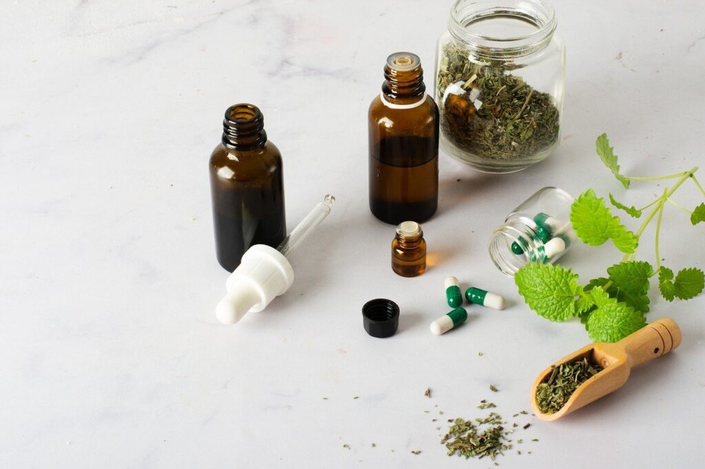 Various glass bottles and jars filled with herbs, capsules, and liquid medicine, alongside a sprig of fresh mint on a white marble surface.