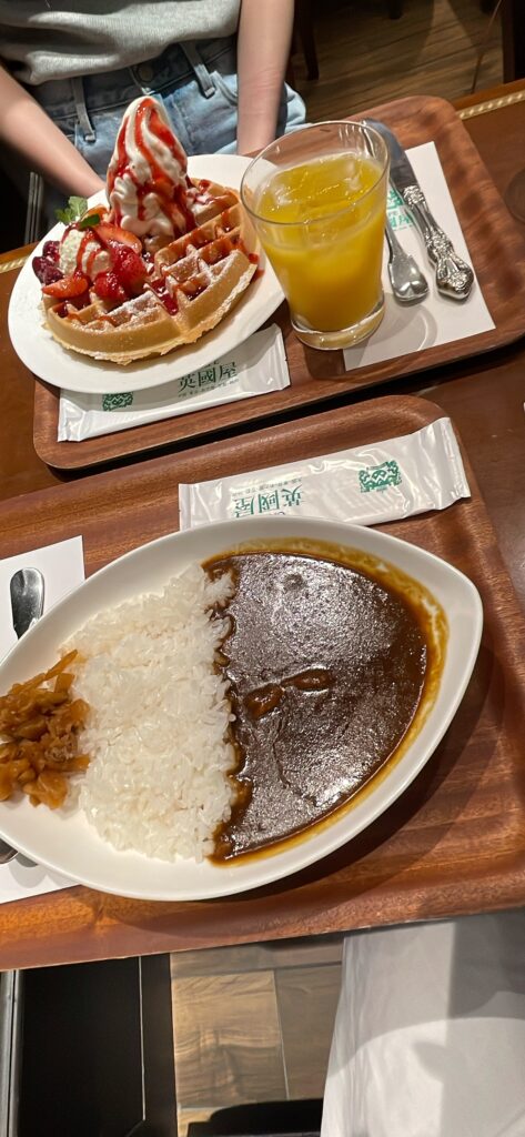 A plate of Japanese curry rice with pickles, alongside a serving of strawberry waffles topped with ice cream and a glass of orange juice in Umeda.