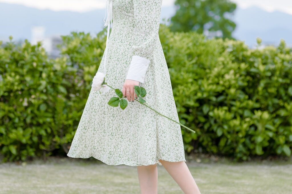 A person in a floral green dress holding a single white rose, walking in a serene garden.