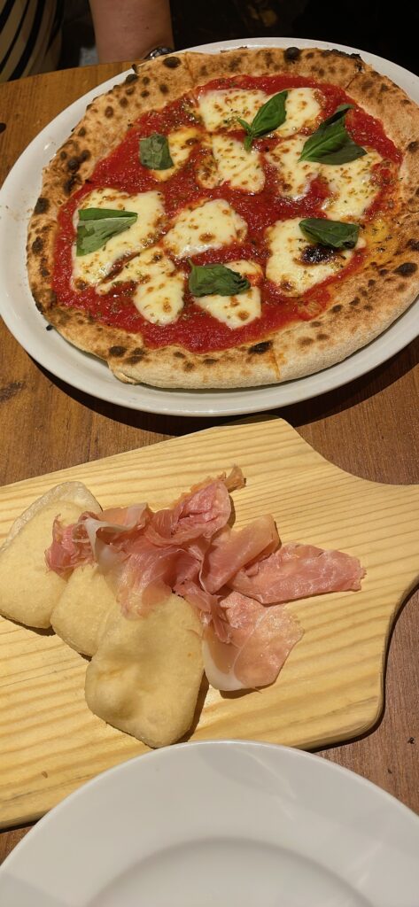 A freshly baked Margherita pizza with basil and cheese, served with an appetizer of prosciutto and fried dough.