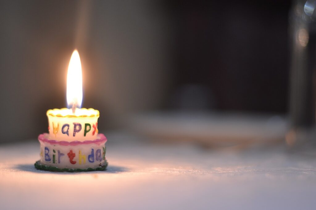 A small tiered birthday cake candle with "Happy Birthday" written on it, glowing in a dimly lit setting.