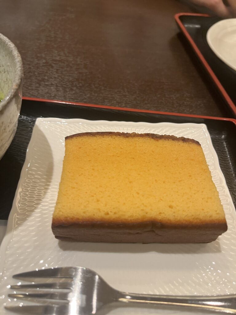 A golden-brown slice of castella sweet sponge cake served on a white plate with a fork, placed on a traditional Japanese tray.