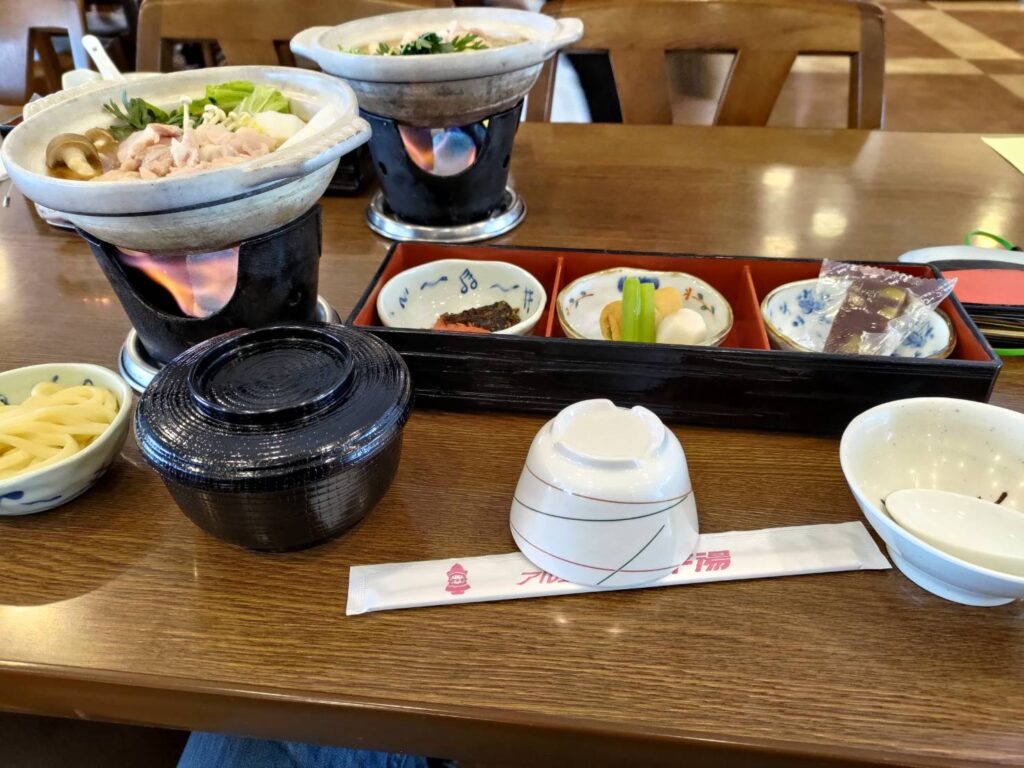 A traditional Japanese hotpot meal served in individual clay pots over open flames, accompanied by a variety of side dishes including pickles, rice, and udon noodles.