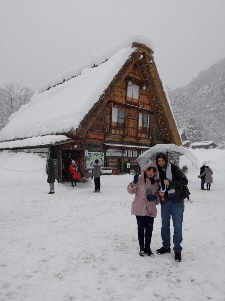  A couple posing in front of a traditional Shirakawago house, smiling under a light snowfall.