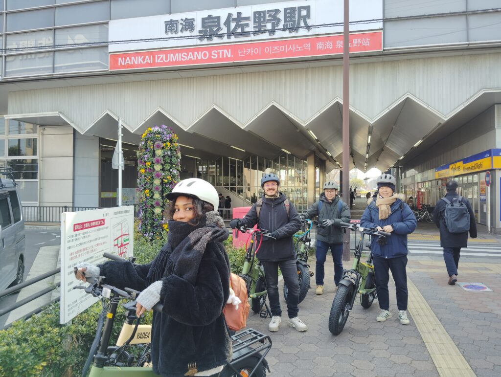 A group of people wearing helmets posing with their e-bikes in front of Nankai Izumisano Station, ready for an adventure in the city.