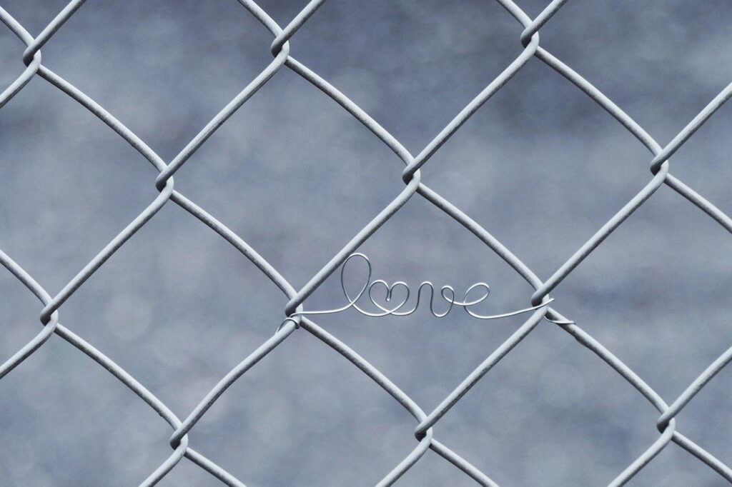 A delicate wire spelling out the word "love" is intertwined with a chain-link fence, symbolizing love’s resilience and presence in unexpected places.