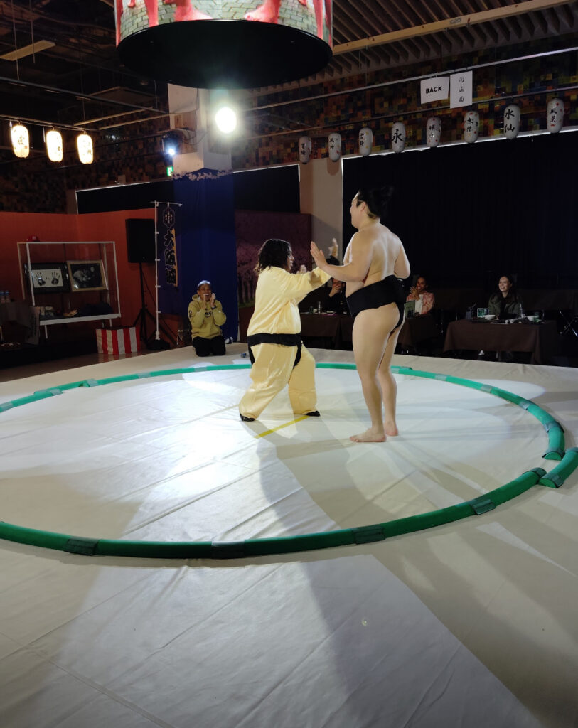 A sumo wrestler and a woman in a sumo suit striking fun poses inside the ring, with traditional lanterns in the background. Ochiizumi