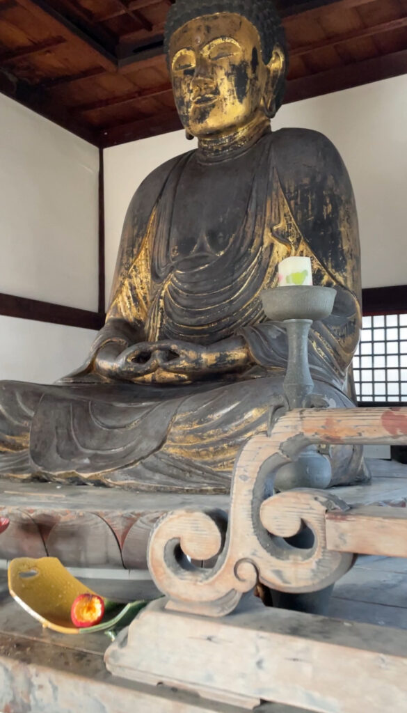 A large, aged Buddha statue with gold accents inside Jozenji Temple, a candle placed in front of it.