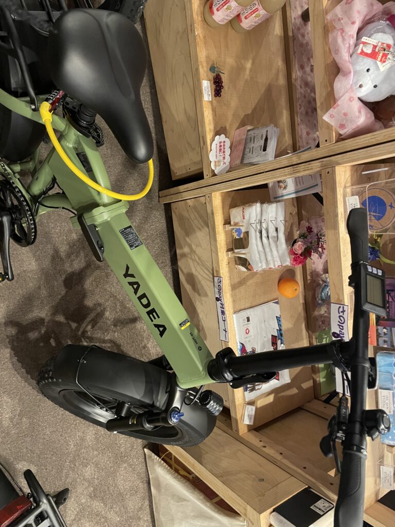 A close-up of a green YADEA electric bike parked indoors, surrounded by wooden shelves with local goods.