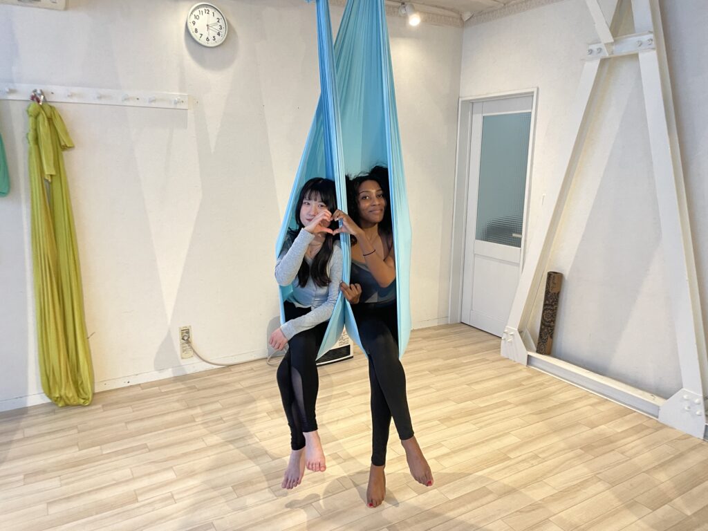 Two friends smiling, sitting comfortably in blue silk yoga hammocks, making a heart with their hands.