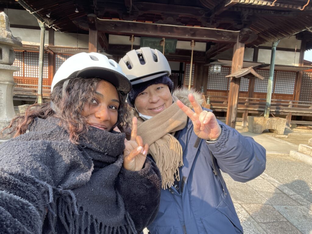 A smiling woman and a tour guide, both wearing helmets, posing for a selfie in front of Jozenji Temple. Ochiizumi