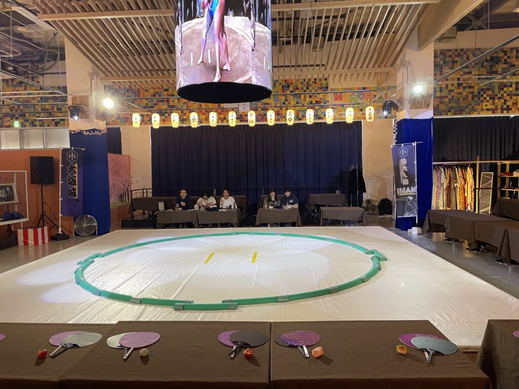 Two sumo wrestlers in traditional robes standing in the sumo ring, introduced on the big screen. A hot pot meal is visible in the foreground.