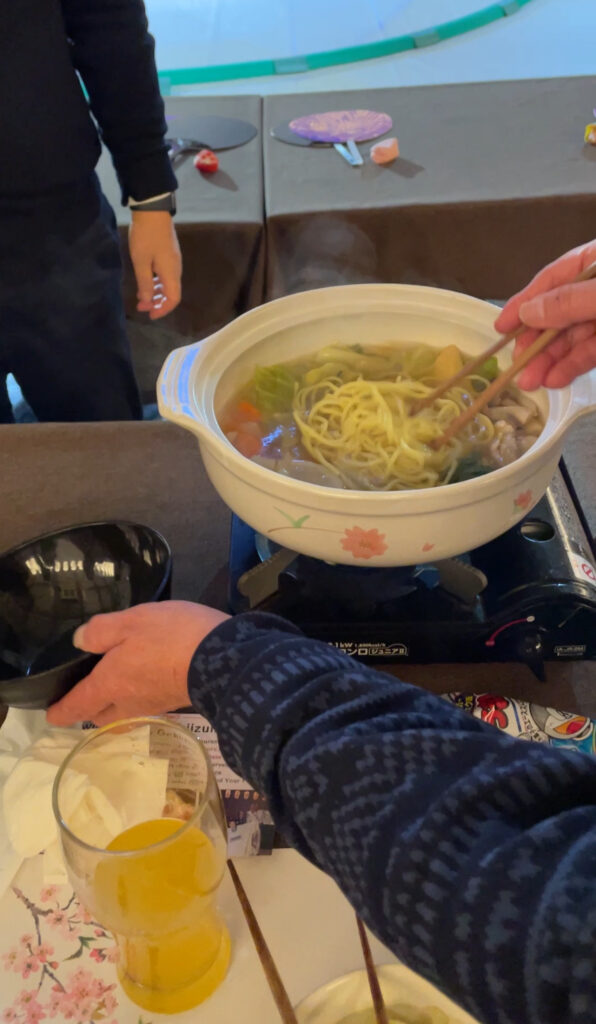 A close-up of a hot pot filled with steaming chanko nabe, with noodles being picked up by chopsticks.