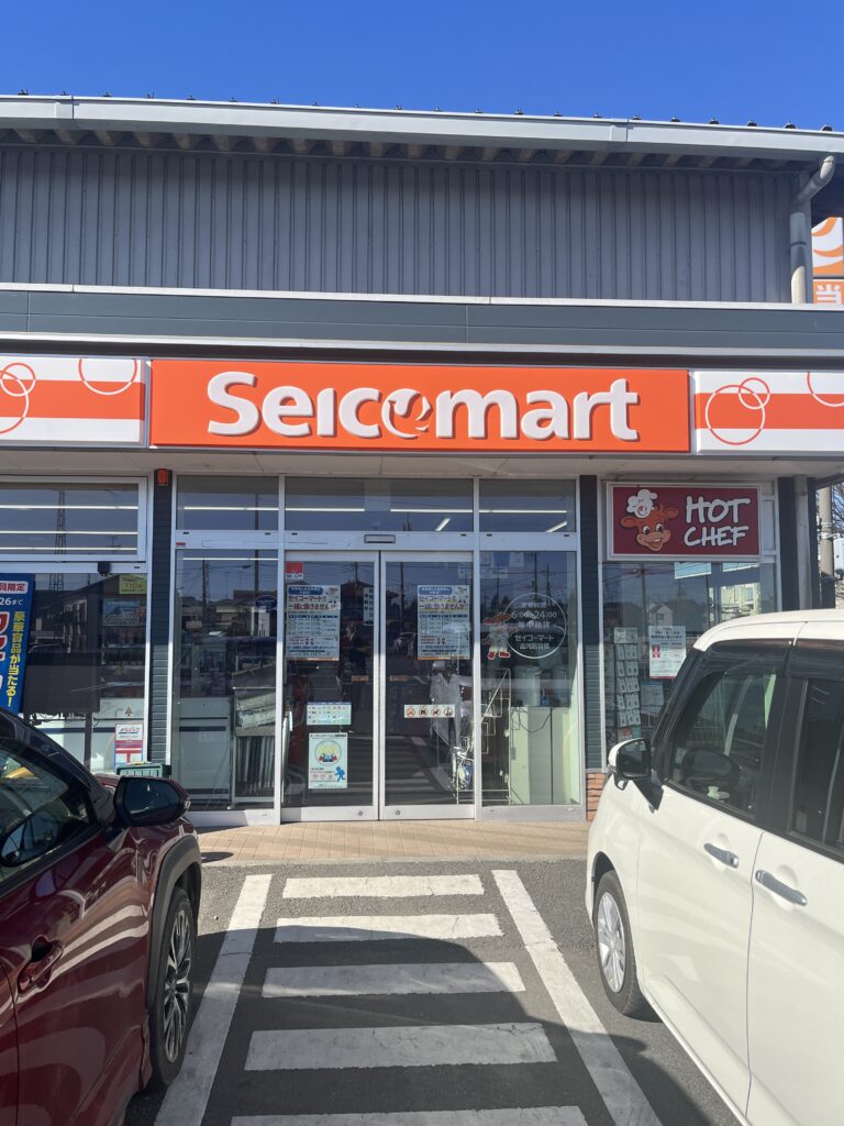 The entrance of a Seicomart convenience store, featuring its signature bright orange signage. The "Hot Chef" section is prominently displayed, advertising fresh and hot food options.