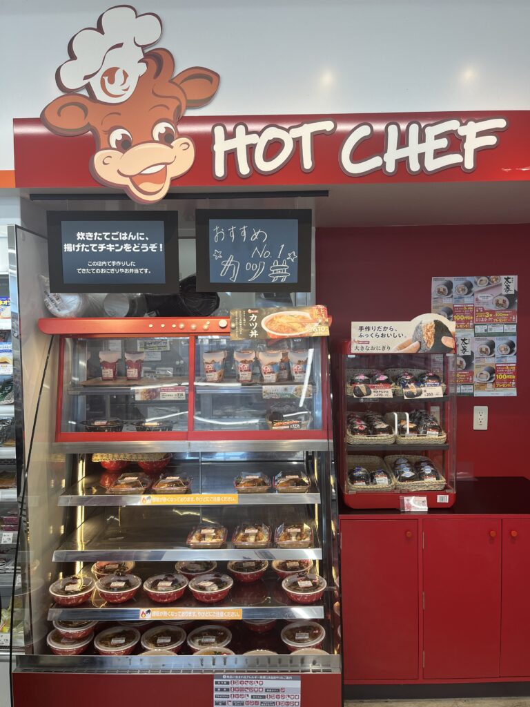 The Hot Chef food counter inside a Seicomart store, offering freshly prepared meals like katsudon, onigiri, and fried food items. A cartoon cow mascot wearing a chef’s hat is featured on the sign in convenience store.