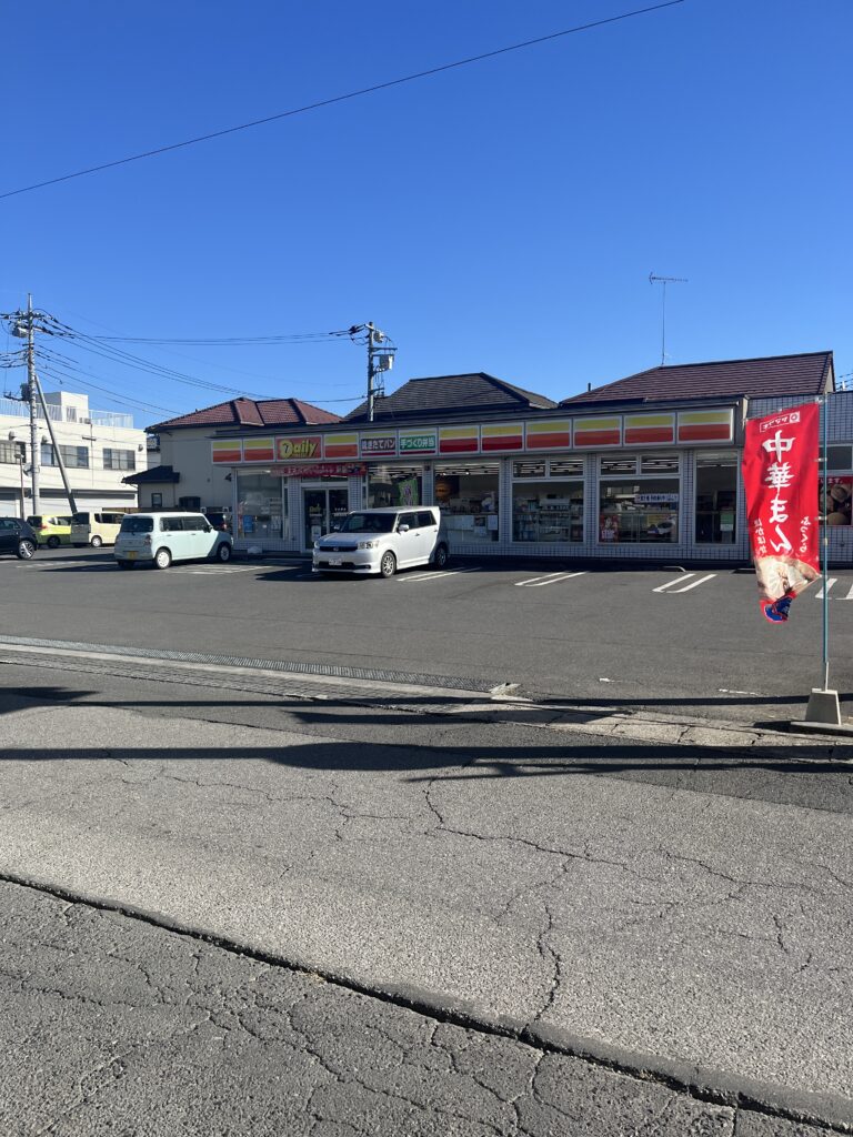 A Daily Yamazaki convenience store in Japan with a parking lot in front. The bright yellow and red signage contrasts with the clear blue sky. A banner advertising Chinese-style buns flutters in the wind.