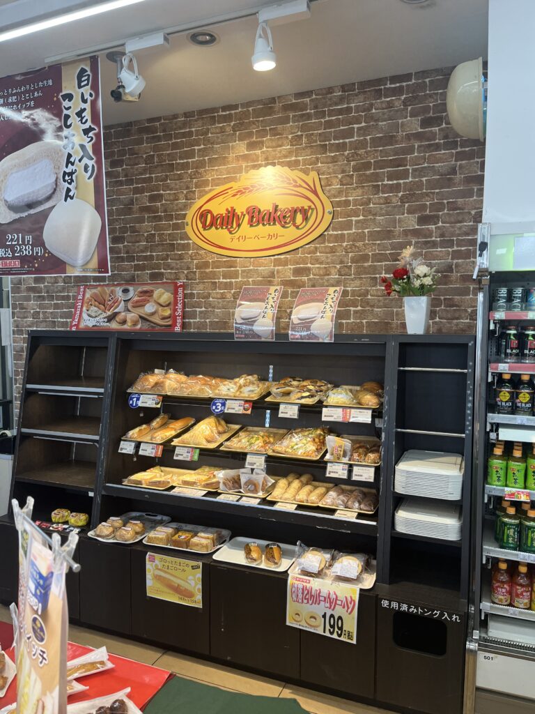 The bakery section inside a Daily Yamazaki convenience store, showcasing an array of freshly baked bread and pastries neatly arranged on shelves. A "Daily Bakery" sign is mounted on a brick-patterned wall.