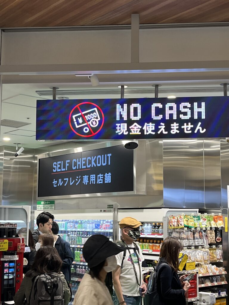 A Newsdays convenience store inside a Japanese train station. The store has a modern, minimalistic design with a digital sign above the entrance displaying "No Cash" and "Self Checkout." Several customers are shopping inside.