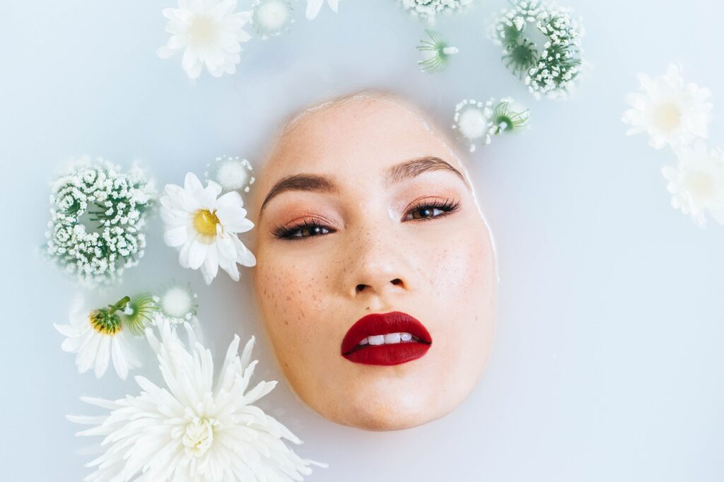 Woman’s face in a floral milk bath with red lipstick and soft makeup.