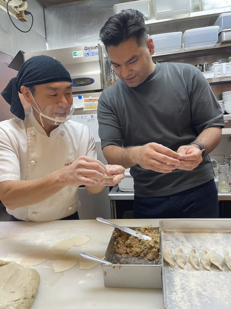 A Japanese chef teaching a man how to make gyoza in a restaurant kitchen.