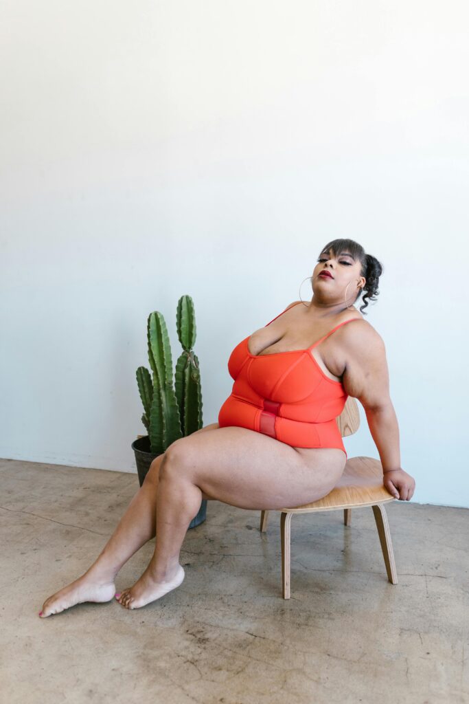 A plus-size woman in a bold red bodysuit sitting confidently on a wooden chair.