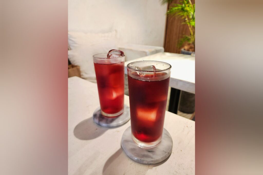 Two glasses of iced cranberry juice on a white table, with a blurred café interior in the background.