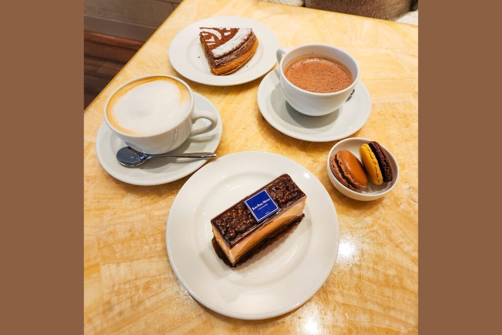 A table set with an elegant dessert spread at Jean-Paul Hevin, including a chocolate cake, macarons, cappuccino, and hot chocolate.