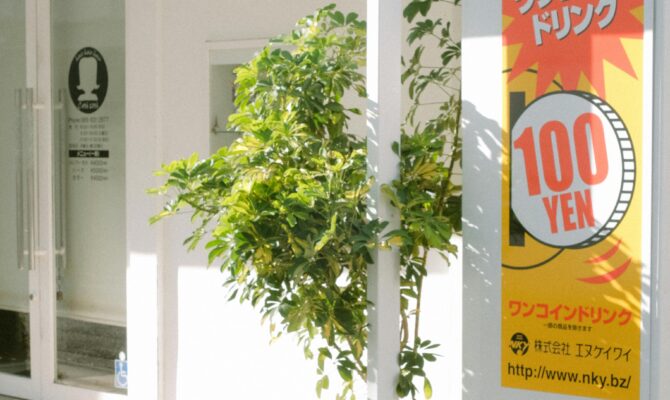 A bright yellow and red sign displaying "100 YEN" outside a white storefront in Japan, surrounded by green plants.