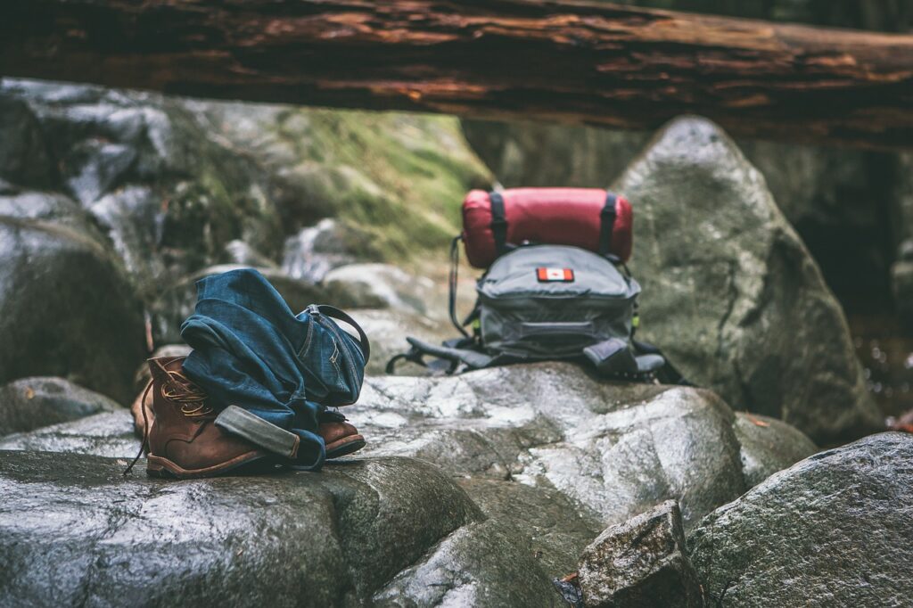 Hiking backpack, rolled-up sleeping mat, boots, and jeans resting on rocks in a forest.