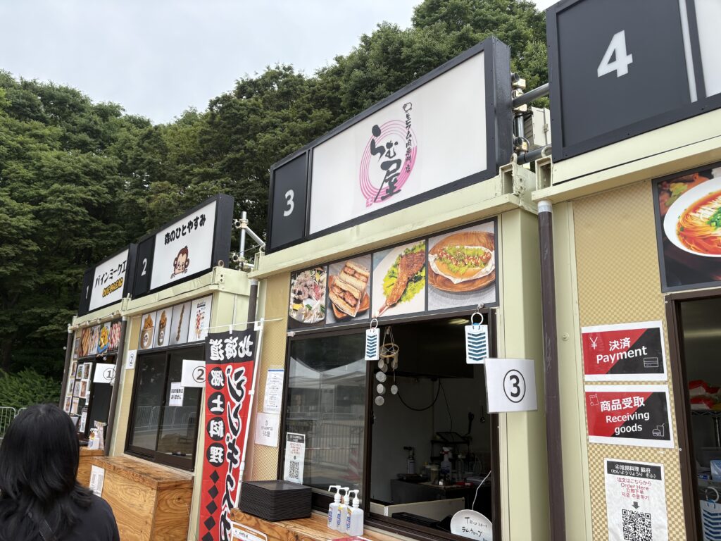 Numbered vendor stalls at the Osaka expo featuring menus with photos and digital payment options.