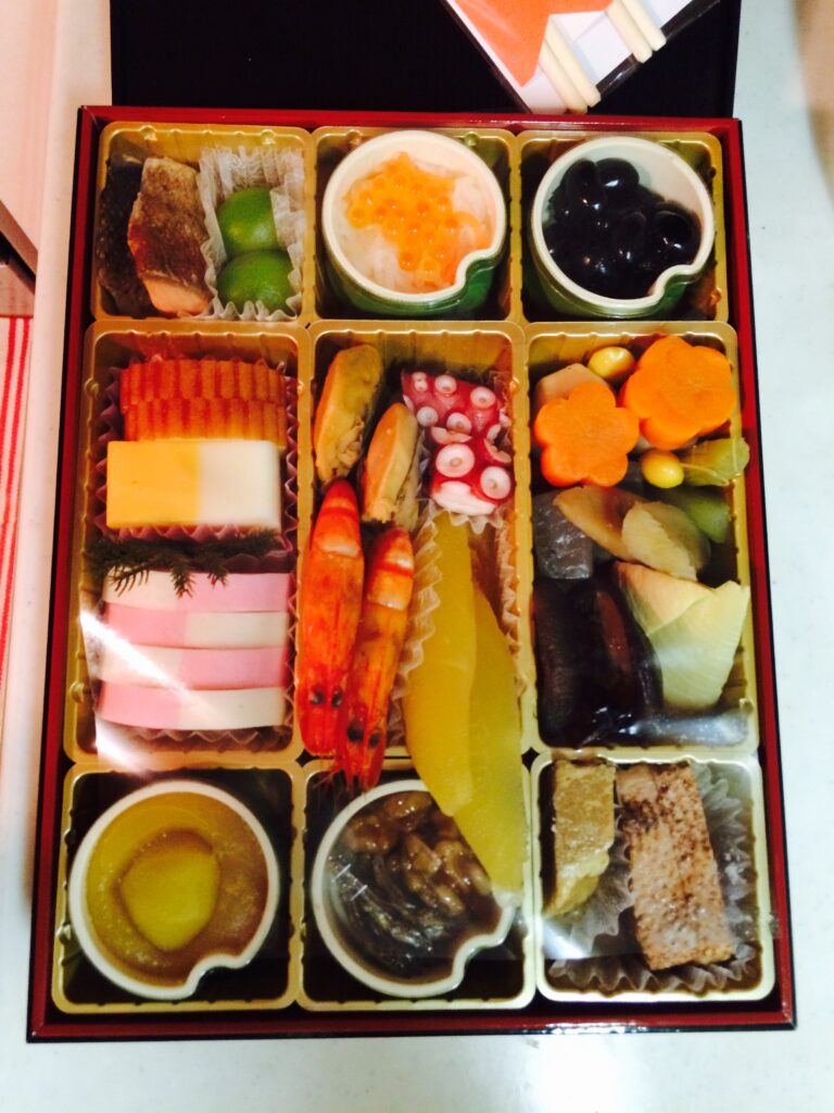 Traditional Japanese New Year osechi bento featuring shrimp, kamaboko, datemaki, kazunoko, black beans, and various simmered vegetables in a lacquered box.