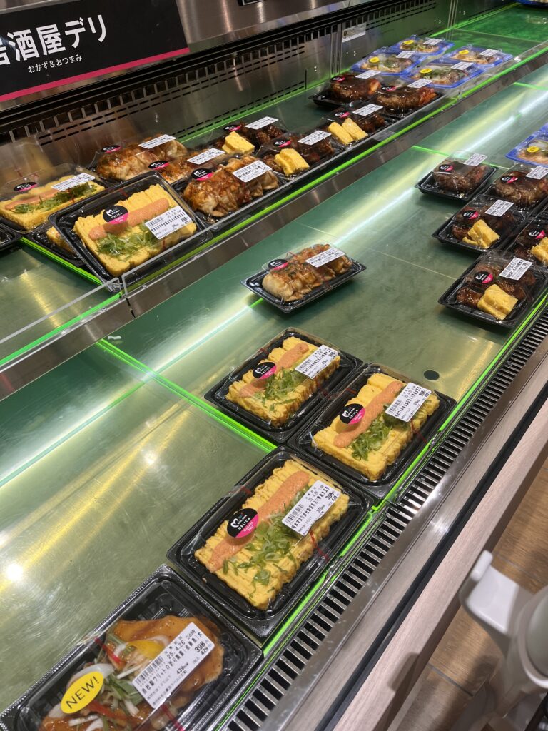 Supermarket deli section with assorted ready-to-eat meals including tamagoyaki topped with mentaiko, yakisoba, and other side dishes neatly arranged in plastic trays.