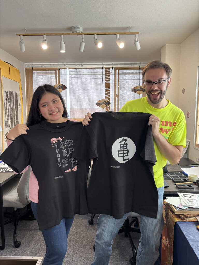 A couple smiling and holding up black t-shirts printed with their completed calligraphy designs at Wa-Pen Calligraphy.