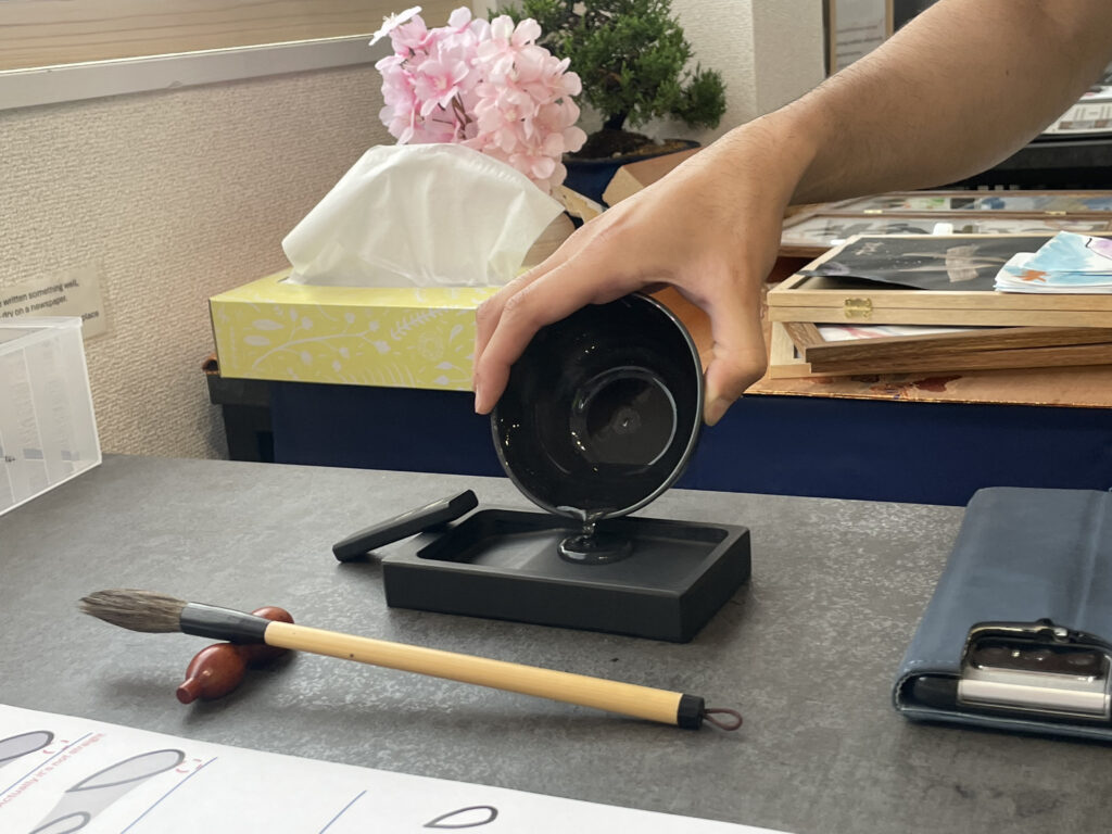 A person pouring black calligraphy ink onto an ink stone, with practice sheets and brush on the desk.