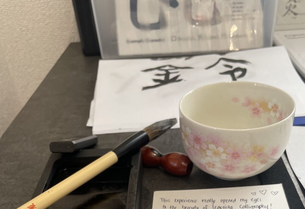 Calligraphy brush, ink stone, sakura-themed tea cup, and finished kanji sheets on a desk at Wa-Pen Calligraphy in Asakusa.