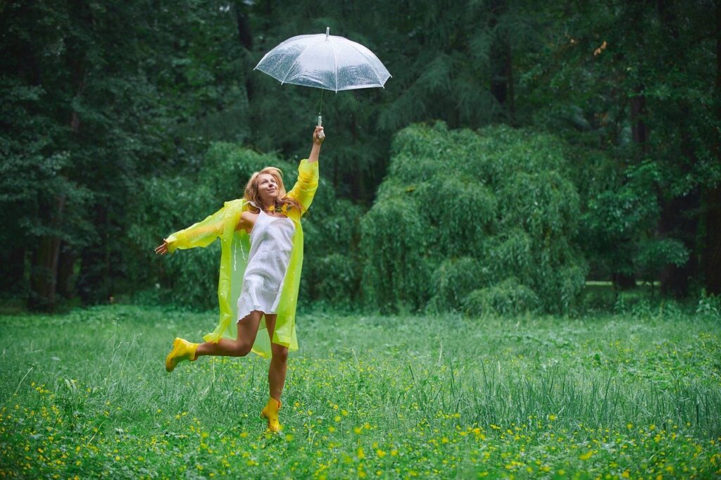 Woman in yellow raincoat jumping happily with umbrella in a green field.
