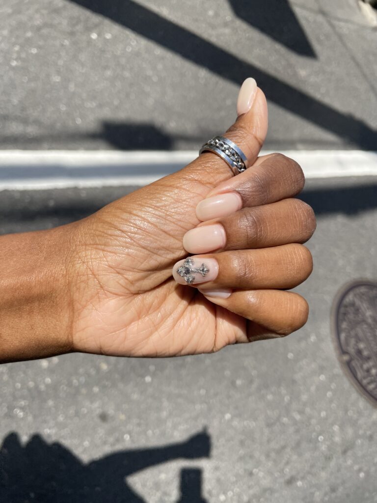 Close-up of a hand with nude gel nails and a sparkly cross design, taken in natural sunlight outdoors.