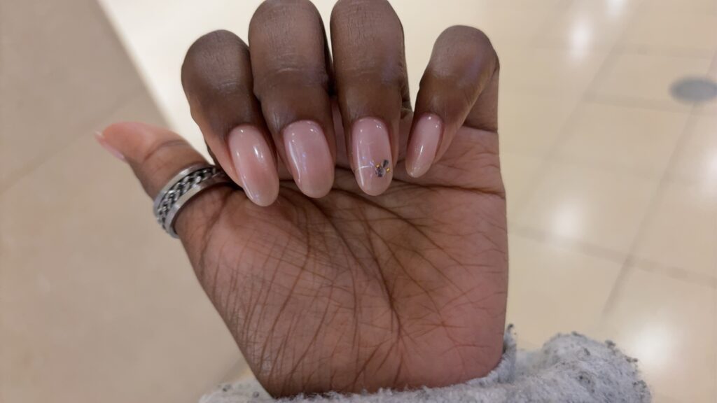 Close-up of a hand with glossy nude pink gel nails and a small floral rhinestone detail, taken indoors.